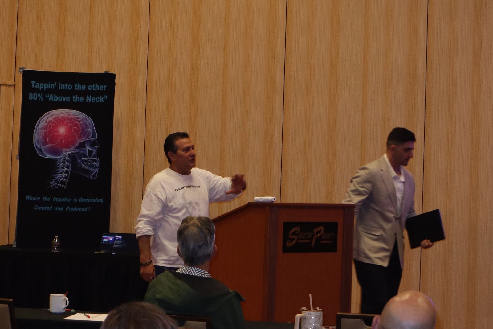 Dr. Adam Del Torto speaking at the South Point podium as Jonathan Tomasian crosses the stage during the seminar's Sunday wrap-up