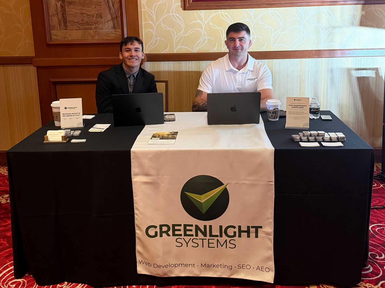 Hunter and Jonathan seated with laptops at the Greenlight Systems booth, behind a table with a Greenlight Systems banner
